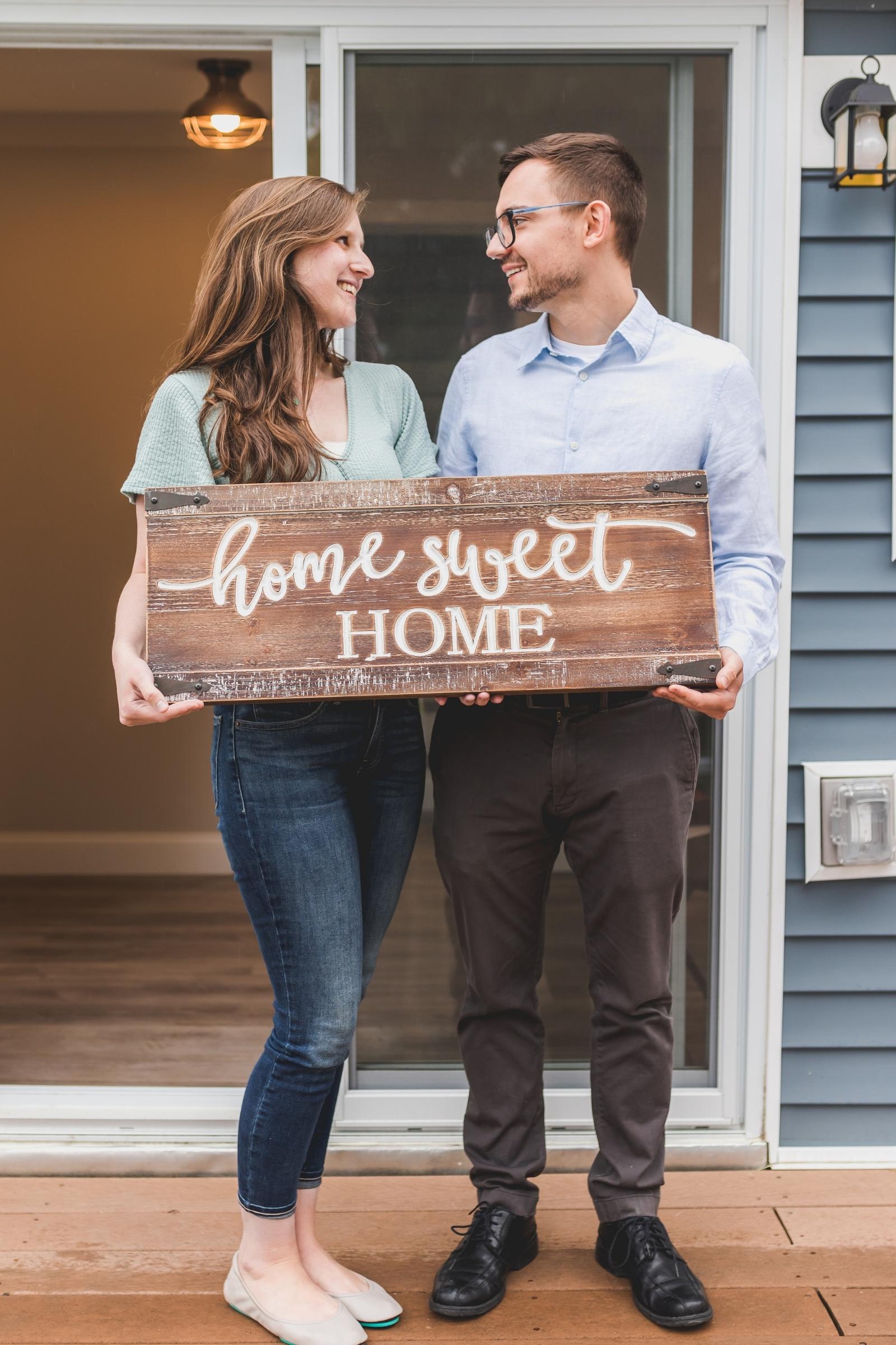 Young couple on a porch holding a wooden 'Home Sweet Home' sign